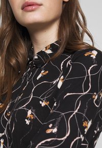 Black blouse featuring a collared neck, floral print in pink and orange, with fluid line patterns on a textured fabric.