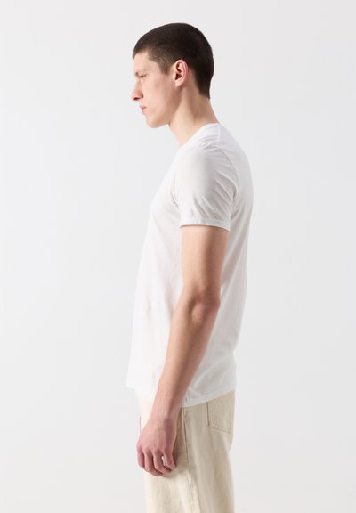Jeune homme debout de profil portant un t-shirt blanc uni à manches courtes et un pantalon beige sur un fond clair.