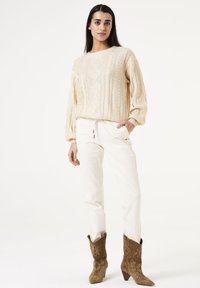 Beige kabelgebreide trui met textuurpatronen, gecombineerd met witte broek en bruine geborduurde cowboylaarzen met een spitse neus.