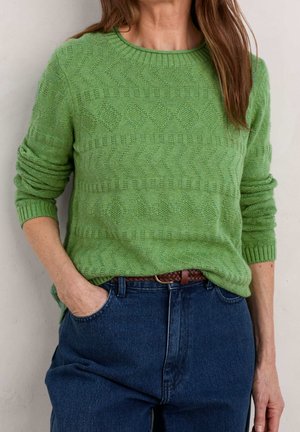Femme portant un pull en maille vert texturé à col rond, rentré dans un jean bleu taille haute avec une ceinture tressée marron.