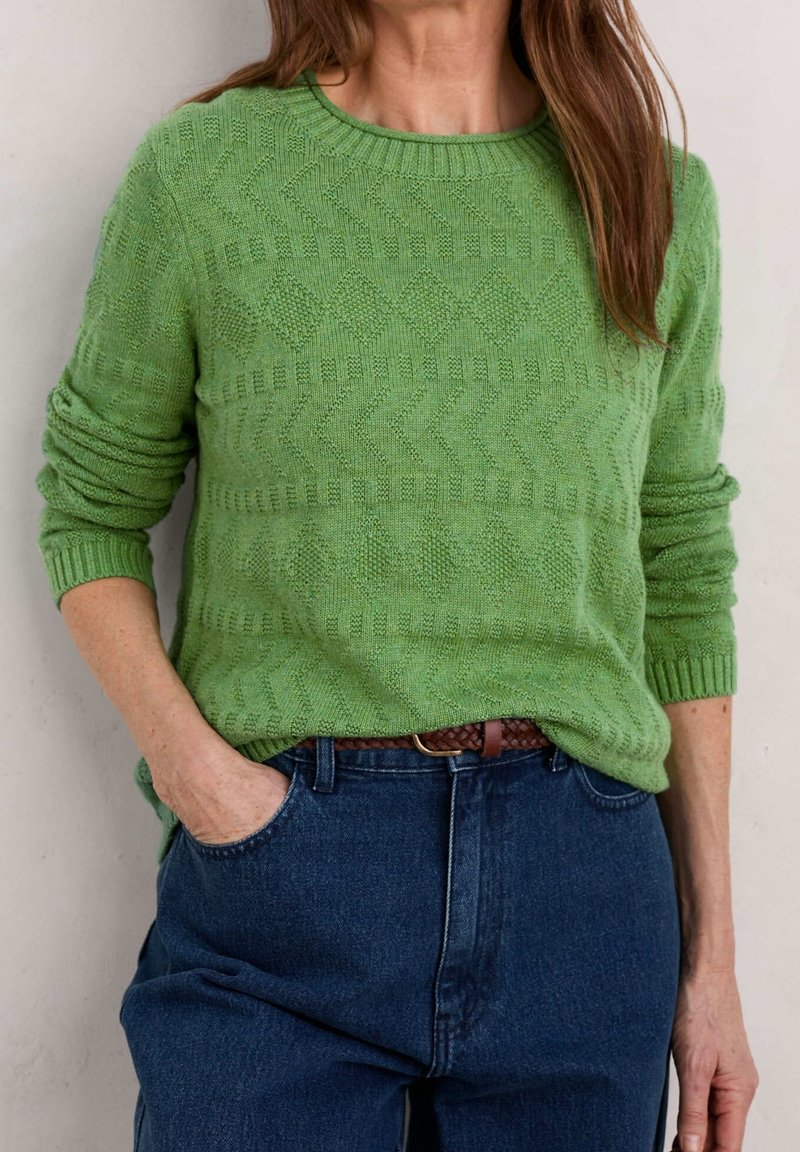 Femme portant un pull en maille vert texturé à col rond, rentré dans un jean bleu taille haute avec une ceinture tressée marron.