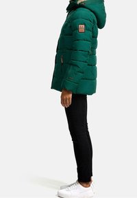 Grön vadderad jacka med huva, med en beige logotyp på ärmen och fickor med dragkedja. Bärs med svarta byxor och vita sneakers.