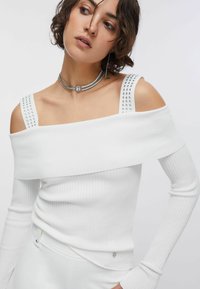 Haut blanc côtelé à épaules dénudées avec manches longues. Présente des clous argentés sur les bretelles et une silhouette ajustée.