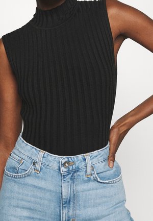 Personne portant un haut noir sans manches à col roulé côtelé, rentré dans un jean en denim bleu clair taille haute, avec la main posée sur la taille.