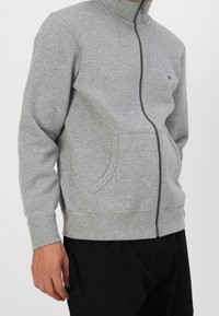 Grauer Kapuzenpullover mit Reißverschluss aus weichem Material, ausgestattet mit Stehkragen, Seitentaschen und gerippten Bündchen. Minimales Logo auf der Brust.