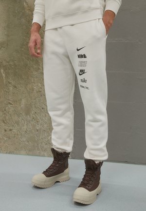 Person trägt weiße Nike-Sweatpants mit mehreren Logo-Stilen am linken Bein und braune High-Top-Stiefel, steht vor gemischtem Wandhintergrund.