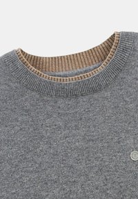 Pull gris avec un col intérieur côtelé marron. Le tissu a une texture douce, et un petit logo est visible en bas à droite.