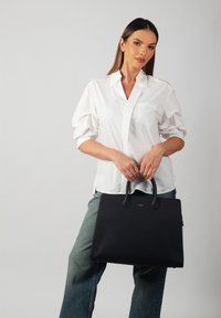 Black leather handbag with textured finish, rectangular shape, and dual handles. Model wearing a white shirt and light denim jeans.