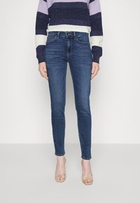 Jeans blue slim fit indossati da una persona in piedi con un maglione a righe blu, bianco e viola e sandali trasparenti con tacco su uno sfondo neutro.