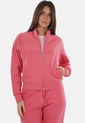 Donna che indossa una giacca rosa con cerniera abbinata e pantaloni, con una mano in tasca, in piedi davanti a uno sfondo bianco semplice.