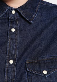 Mörkblå denimskjorta med spetsig krage, pärlknappar, framficka och synliga sömnadsdetaljer. Slät textur, avslappnad design.