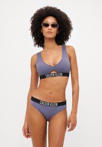 Vijolična rebrasta bikinija seta, ki vključuje zgornji del s izrezom in hlačke visoko pasu, oboje pa ima črno elastično bandico s Calvin Klein logotipom.