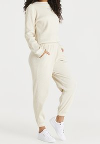 Conjunto de loungewear de color crema con canalé, mangas largas, cinturilla elástica y bolsillos. Combinado con zapatillas blancas para un look casual.