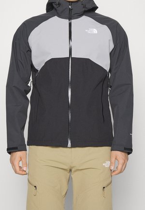Mężczyzna ubrany w czarno-szarą kurtkę z kapturem North Face zapinaną na zamek oraz beżowe spodnie North Face z kieszeniami na zamek.
