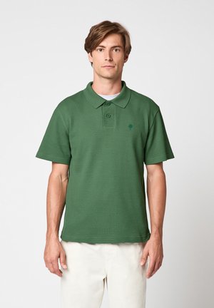 Man draagt een groene polo met korte mouwen en een witte broek, staat tegen een effen lichte achtergrond en kijkt recht vooruit.