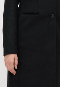 Μαύρο μάλλινο παλτό με κομψή γραμμή, διαθέτοντας κλείσιμο με ένα κουμπί, πλευρικές τσέπες και απαλό υφή.