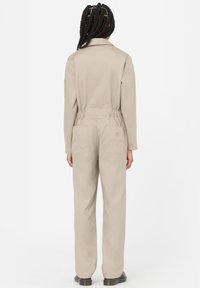 Mono beige con mangas largas, cinturilla elástica, cuatro bolsillos y un cuello. Tejido suave con un acabado mate y un corte relajado.