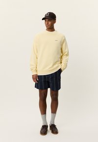 Κίτρινο φούτερ με στρογγυλή λαιμόκοψη, ναυτικές ριγέ σορτς, γκρι κάλτσες, καφέ παπούτσια, μαύρο καπέλο με τον αριθμό 11, στέκεται σε ουδέτερο φόντο.