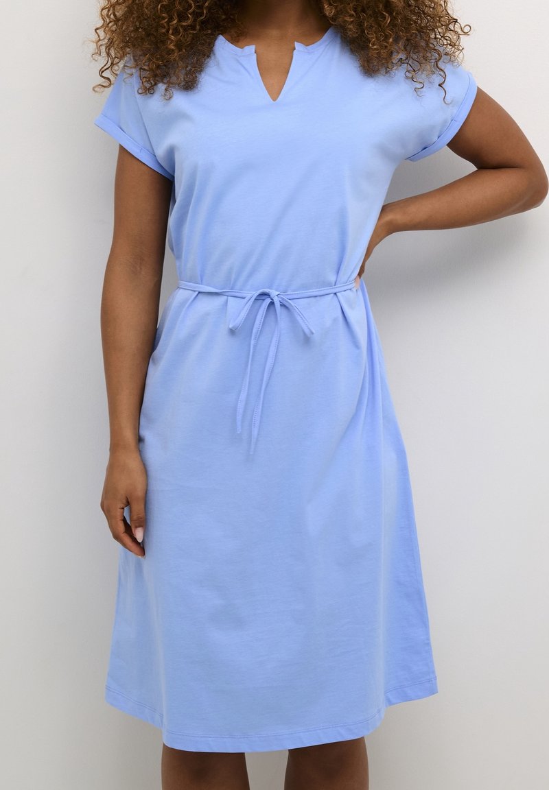 Femme portant une robe bleu clair jusqu'aux genoux, manches retroussées et ceinture nouée à la taille, debout avec une main sur la hanche.