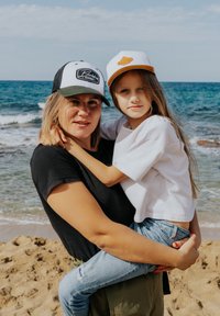 Zwei Personen am Strand, eine trägt ein schwarzes T-Shirt und eine schwarz-grüne Kappe, die andere ein weißes Hemd und eine Kappe mit einem braunen Akzent.