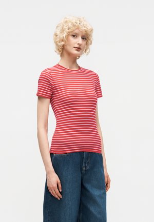 Ξανθιά γυναίκα με σγουρά μαλλιά που φοράει κόκκινο και άσπρο ριγέ κοντομάνικο πουκάμισο και φαρδιά μπλε τζιν, στέκεται μπροστά σε λευκό φόντο.