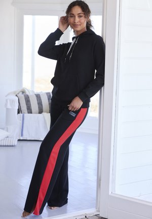 Frau lehnt an einem weißen Türrahmen, trägt einen schwarzen Hoodie und schwarze Hosen mit einem breiten roten Streifen, steht barfuß in einem hellen Raum.