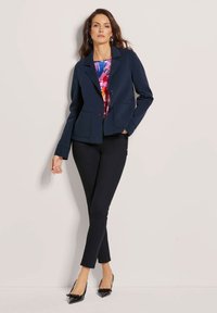 Marineblazer mit zwei Vordertaschen, einreihiger Knopfverschluss und taillierter Passform, kombiniert mit einem bunten Blumenshirt und schmalen schwarzen Hosen.