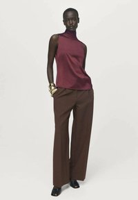 Burgundska brezrokavnik s turtleneck ovratnikom v kombinaciji s svetlo rjavimi širokimi hlačami. Model nosi zlate zapestnice in črne odprte čevlje.