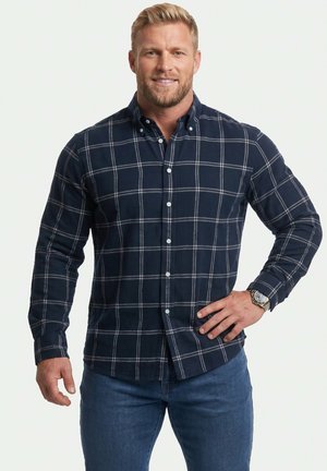 Mand med blond hår iført en marineblå ternet skjorte med knapper og blå jeans, står med den ene hånd på hoften mod en hvid baggrund.