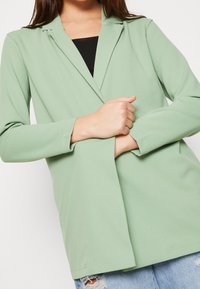 Ljusgrön blazer med långa ärmar, slagkrage, slätt tyg, enknappslåsning, bärs över en svart topp, tillsammans med slitna jeans.