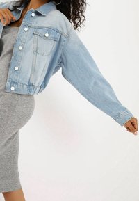 Light blue denim cropped jacket with dropped shoulders, button closures, and a front pocket. Worn over a gray ribbed dress.