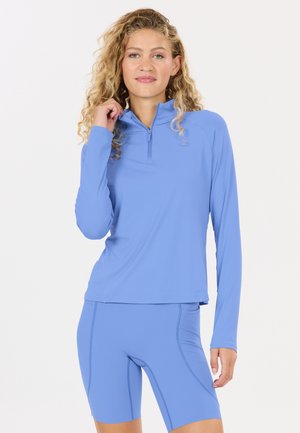 Vrouw met krullend blond haar, gekleed in een lichtblauw langwerpig shirt met rits en bijpassende fietsbroek, staand tegen een effen lichte achtergrond.