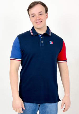 Jeune homme souriant, portant un polo bleu marine avec une manche gauche bleue, une manche droite rouge, col et bords de manches rayés.