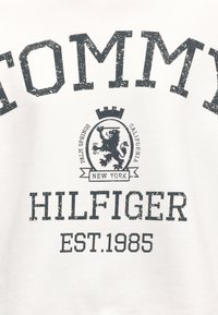 Μπλουζάκι με φθαρμένο κείμενο «Tommy Hilfiger EST. 1985» και ένα έμβλημα με λιοντάρι, κορώνα και τοποθεσίες Palm Springs, Καλιφόρνια, Νέα Υόρκη.