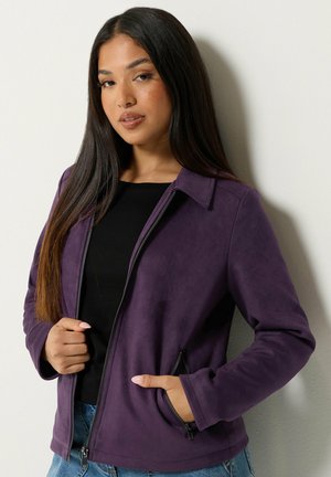 Jeune femme aux longs cheveux foncés portant une veste zippée violette sur un haut noir et un jean bleu, debout contre un mur clair.