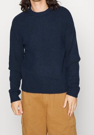 Person trägt einen marineblauen, langärmeligen Strickpullover und hellbraune Cargohosen, die Hände locker an den Seiten.