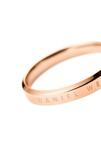 Ruusu-kulta metalli käsirengas, jossa on sileä pinta ja kaiverrus "DANIEL WELLINGTON" yhdellä puolella. Ohut, tyylikäs muotoilu.