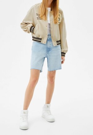 Femme portant une veste bomber beige avec des rayures noires, un short en jean bleu clair, des baskets blanches, debout sur fond blanc.