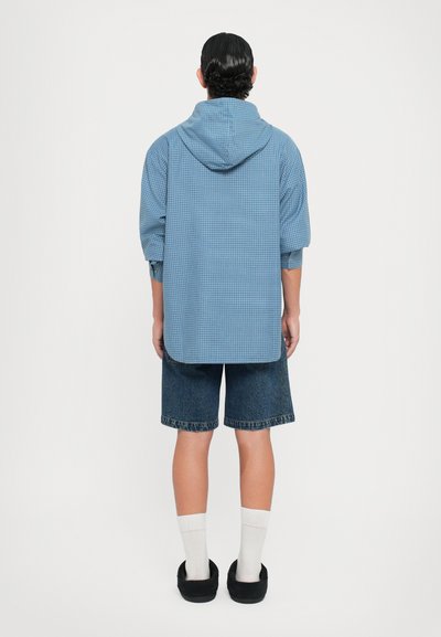 Camisa de cuadros azul con capucha, mangas cortas y dobladillo curvado, combinada con pantalones cortos de denim y zapatillas negras sin cordones, con calcetines blancos visibles.