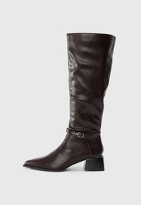 Botte en cuir marron foncé, haute, à bout pointu, avec une petite sangle à boucle autour de la cheville et un petit talon carré sur fond blanc.