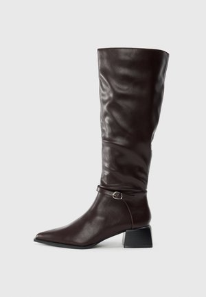 Bota alta de cuero marrón oscuro con punta afilada, pequeña correa con hebilla alrededor del tobillo y tacón bajo en fondo blanco.