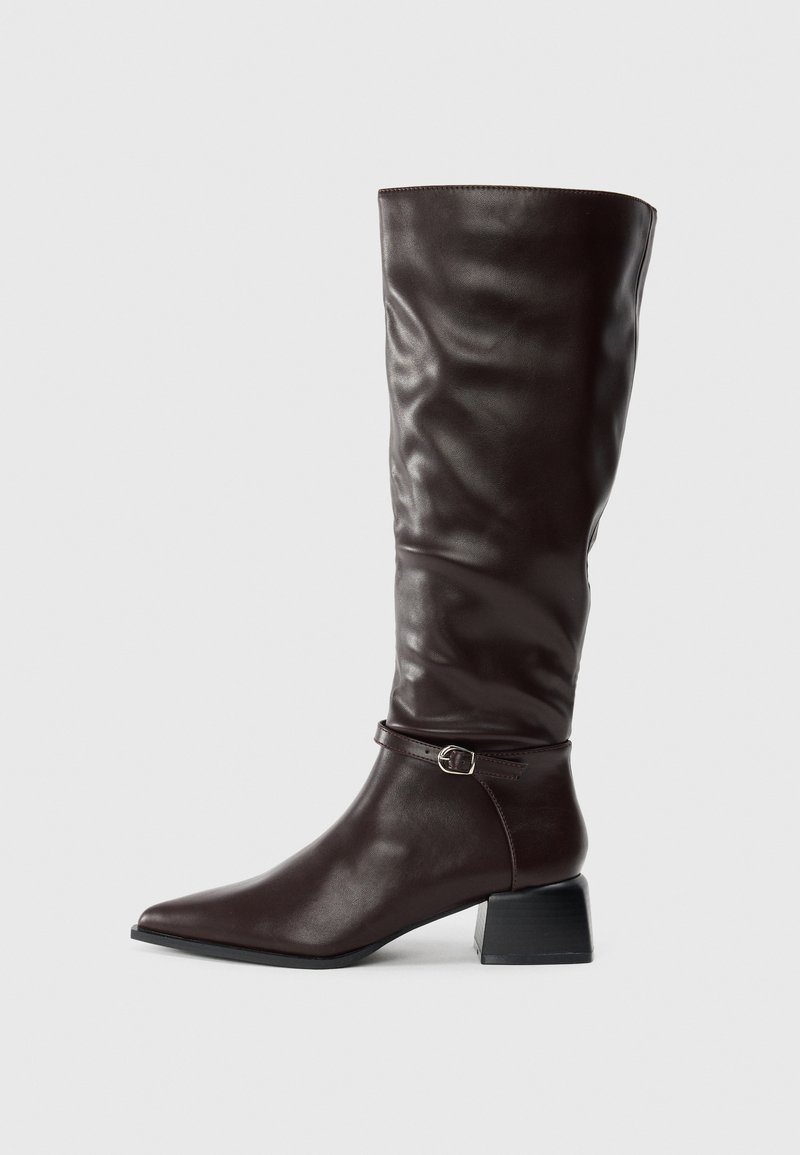 Botte en cuir marron foncé, haute, à bout pointu, avec une petite sangle à boucle autour de la cheville et un petit talon carré sur fond blanc.