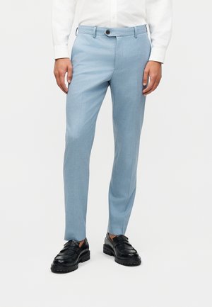 Man wearing light blue tailored trousers, white shirt, and black leather loafers, standing against a plain white background.