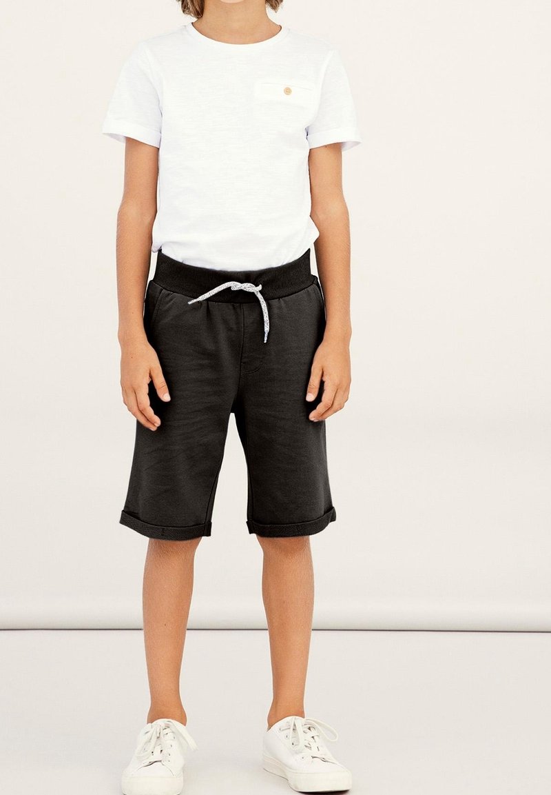 Enfant portant une chemise blanche à manches courtes, un short noir avec cordon et des baskets blanches, se tenant devant un fond clair uni.