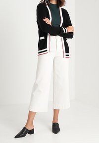 Svart cardigan med röda och vita ränder, matchad med vita vida jeans och svarta klackskor. Cardiganen har fickor och knappar.