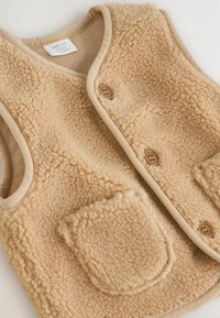 Gilet in pile beige con una superficie morbida e texture, dotato di tasche arrotondate, bordi color nocciola e due bottoni frontali. Design leggero e accogliente.