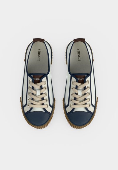 Ζευγάρι λευκά και ναυτικά μπλε sneakers Versace με μπεζ κορδόνια και καφέ λαστιχένιες σόλες, εμφανιζόμενα από πάνω σε λευκό φόντο.