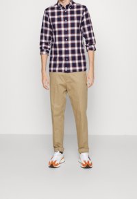 Camicia button-up da uomo con un motivo a quadri blu navy, rosso e bianco, abbinata a pantaloni beige e sneakers bianche con accenti arancioni e neri.