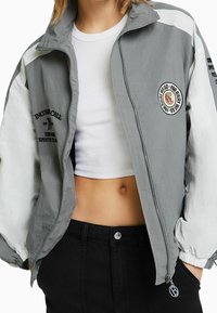 Šedá a bílá bunda na zip s vysokým límcem, elastickými manžetami a grafickými nášivkami. Materiál z bavlněné směsi, zkrácený střih a lehká textura.