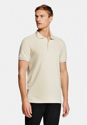 Polo beige en tissu texturé avec col et manches courtes, associé à un pantalon noir. Design épuré et minimaliste.